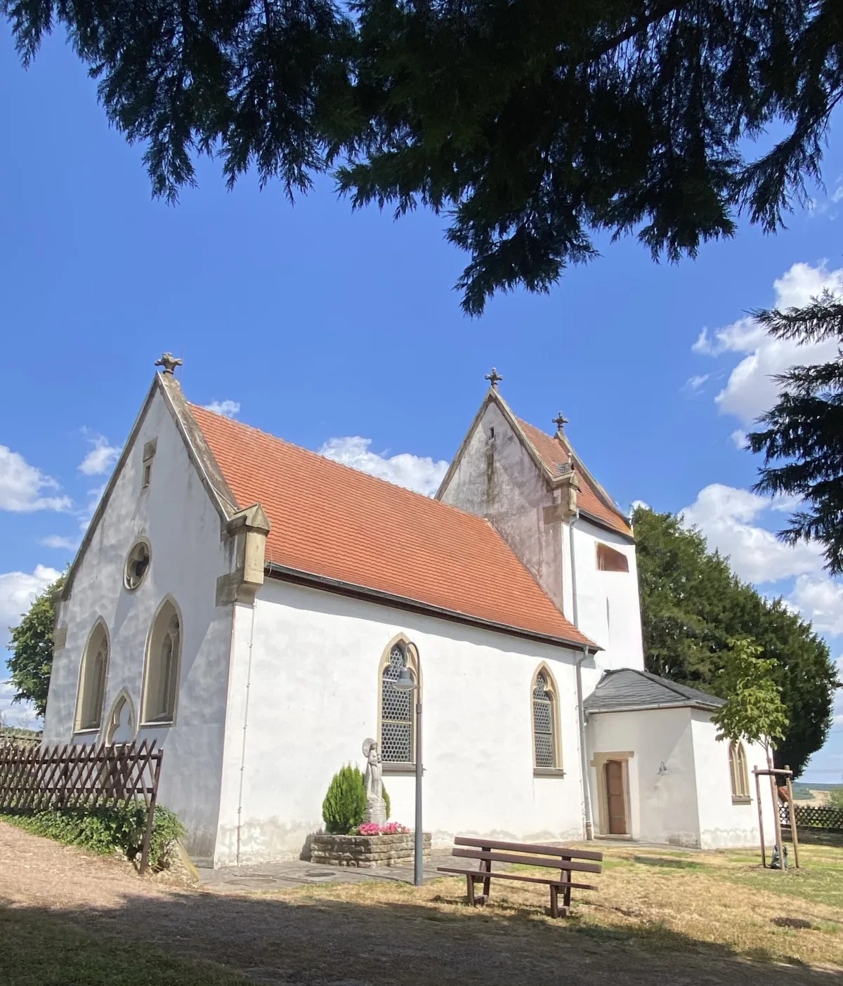 Taufkirche Hildegards von Bingen in Bermersheim vor der H&ouml;he: wei&szlig; verputzter Bau mit rotem Ziegeldach und gotischen Fenstern, davor eine Bank unter B&auml;umen