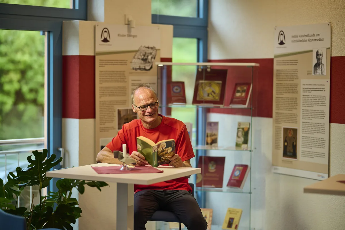 Besucher liest ein Hildegard-Buch in der Begegnungsst&auml;tte am Hildegard-Pilgerwanderweg, dahinter Ausstellungstafeln und eine Vitrine mit B&uuml;chern