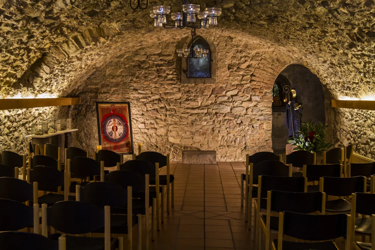 Beleuchteter historischer Gew&ouml;lbekeller mit Stuhlreihen, einer reproduzierten Hildegard-Miniatur an der Bruchsteinwand und einer Hildegard-Figur in einer Nische