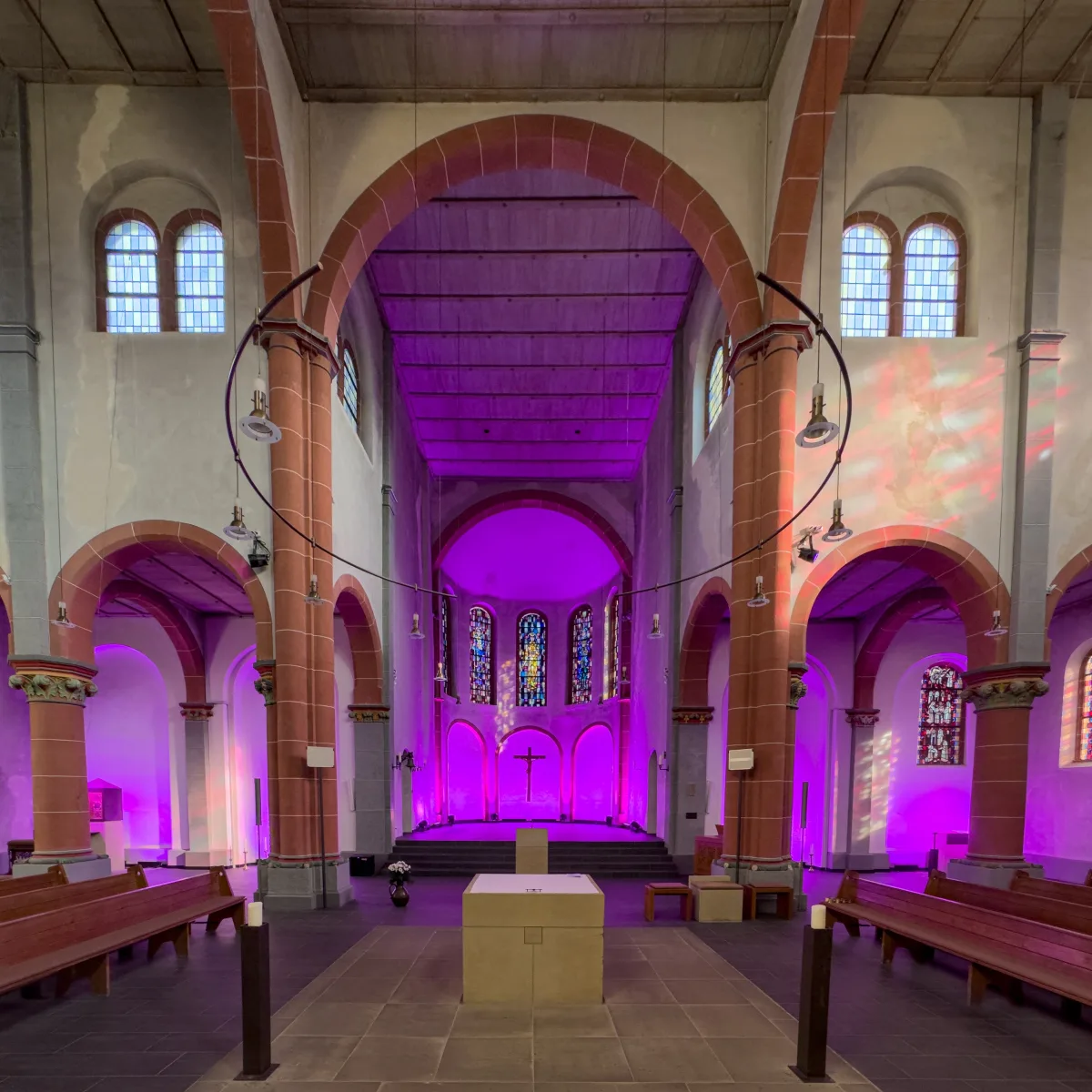 Romanischer Kirchenraum des Hildegard-Zentrums in violettes Licht getaucht, Blick durch das Mittelschiff auf den Altarraum mit Kreuz